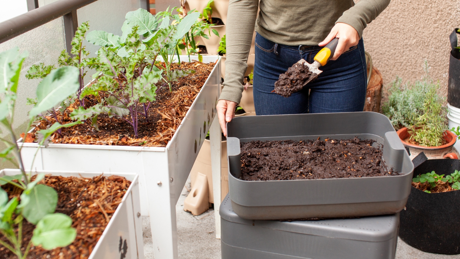 A woman stands on her garden balcony, holding a trowel filled with a small pile of dark worm castings.