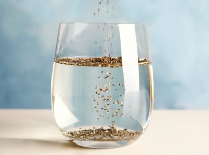 Seeds sink or float test. Pouring chia seeds into glass with water on a wooden table, on a blue background. Some seeds float while others remain at the bottom of the glass. The seeds are small, round in shape, and black in color.