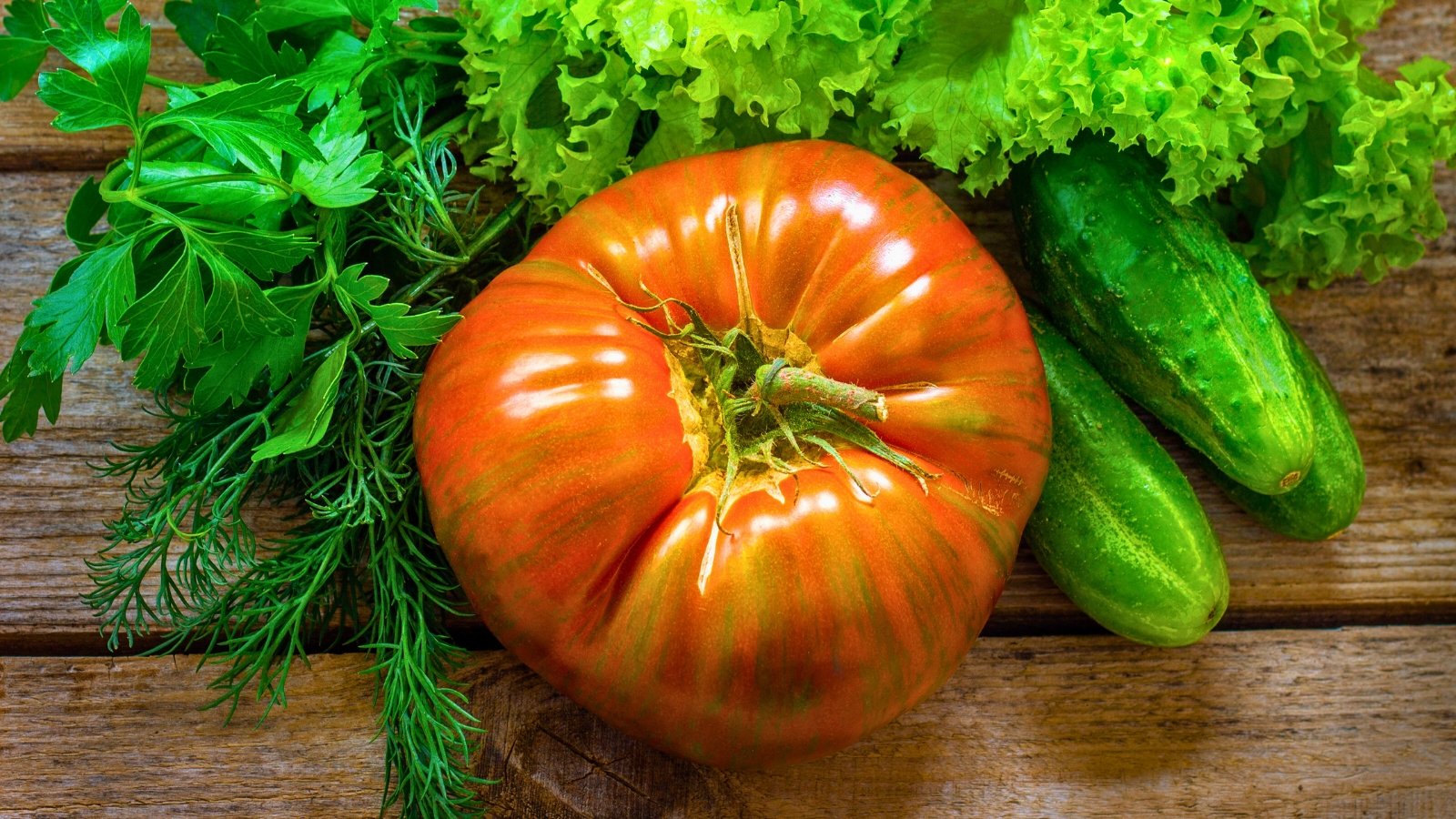 An orange Berkeley Tie-Dye tomato sits alongside fragrant rosemary, fresh parsley, crisp lettuce, and crunchy cucumbers atop a rustic wooden table.