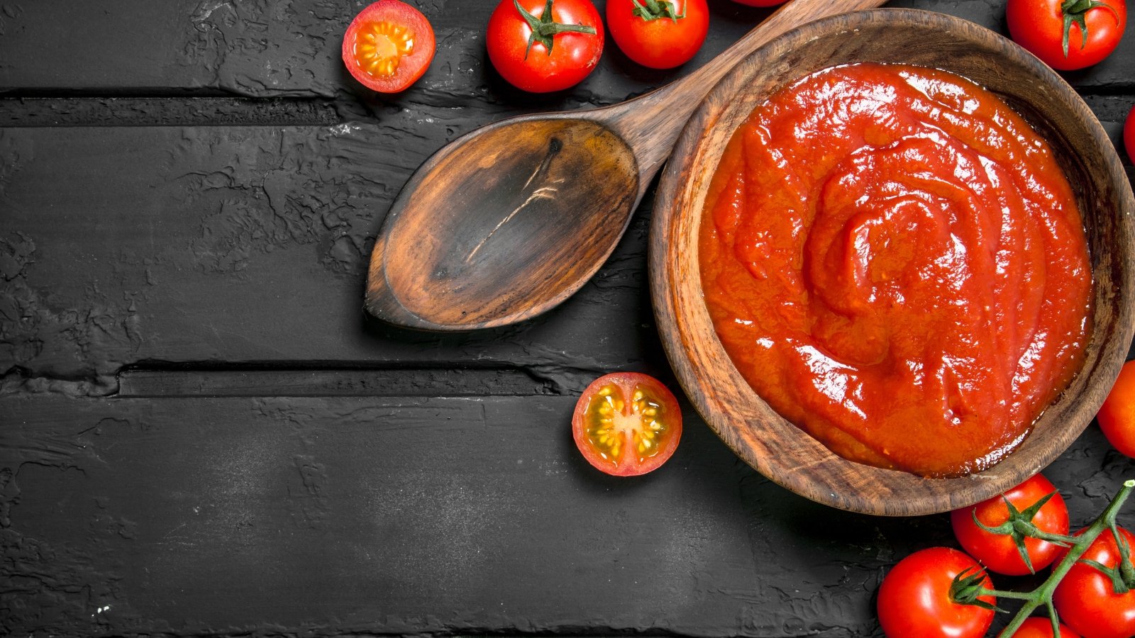 Top view, close-up of tomato sauce in a wooden bowl next to a wooden spoon and several fresh cherry tomatoes on a black wooden surface.